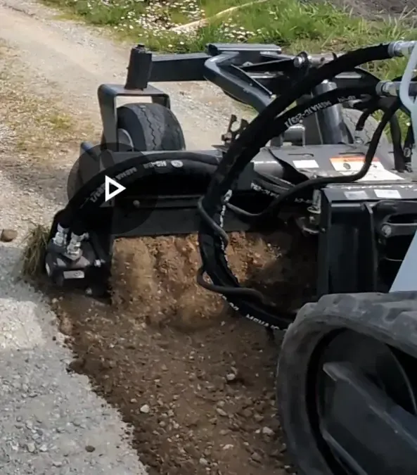 A soil tiller machine is actively working on a patch of dirt, breaking up the soil. The machine is on a gravel path with some greenery visible in the background.